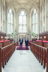 Two men in formal suits stand at the front of an ornate, grand hall with high arched windows, wooden pews, stained glass, and floral arrangements, suggesting a wedding or ceremonial event.