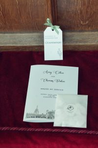 A wedding order of service booklet, a white tag labelled This seat is reserved for Groomsman with a green ribbon, and a small white envelope, all placed on a red velvet surface.