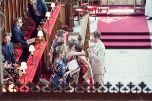 A group of people in formal attire greet and hug each other inside a church with wooden pews and red cushions; one man in a light suit stands nearby holding a piece of paper.