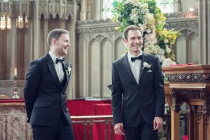 Two men in dinner jackets smile and stand together at the front of a church decorated with flowers and candles, suggesting they are at a wedding ceremony. Ornate stone arches and stained glass windows are in the background.