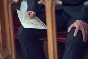A person in a black suit sits on a red cushioned bench, holding a folded programme or booklet with handwritten text, inside a wooden-panelled setting, possibly a church or formal event.