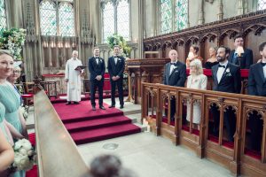 A wedding ceremony in a church shows two grooms in tuxedos standing together at the altar, surrounded by wedding guests and an officiant in white robes. The guests watch from wooden pews. The church has large stained glass windows.