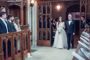A bride in a white gown, holding a bouquet, walks down the aisle on the arm of an older man in a dinner jacket. Guests seated on wooden pews watch them in a church with ornate wooden panelling.