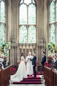 A bride and groom stand before a priest in a grand church with tall stained glass windows, exchanging vows as guests watch from wooden pews decorated with white flowers.