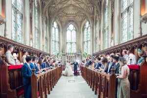 A bride and groom walk down the aisle of a grand, ornate hall with high ceilings and stained glass windows, as guests seated on both sides watch and take photos.
