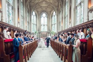 A bride and groom walk down the aisle together in a grand, ornate church as guests seated in wooden pews on either side watch and smile. Sunlight streams through tall stained glass windows in the background.