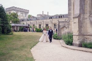 A bride and groom walk hand in hand along a gravel path outside a historic stone building, with wedding guests gathered under a wooden shelter in the background.