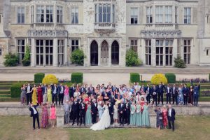 A large wedding group poses happily outside a grand, historic manor. The bride, groom, and wedding party stand at the front, with guests in formal attire arranged in rows behind them on stone steps.
