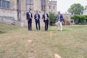 Four men dressed in suits and sunglasses play an outdoor lawn game with wooden blocks on a grassy area near a historic stone building.