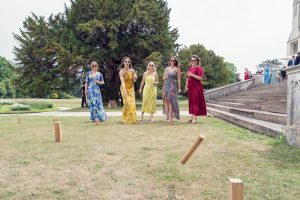 Five women in colourful dresses walk across a grassy lawn while playing an outdoor game with wooden blocks, near stone steps and a large tree, with more people and a building in the background.