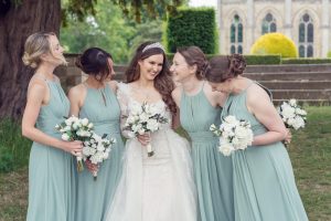 A bride in a white wedding dress and tiara stands outdoors, smiling and holding a bouquet, surrounded by four bridesmaids in matching light blue dresses, each holding white bouquets. They are laughing together. A church is in the background.