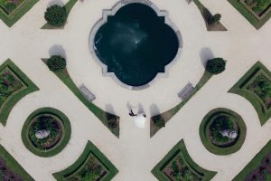 Aerial view of a bride and groom walking near a decorative garden fountain, surrounded by symmetrical paths, green shrubs, and manicured flowerbeds. Their shadows are visible on the pale gravel path.