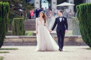 A bride in a white, long-sleeved gown and a groom in a black tuxedo walk hand in hand outdoors, smiling at each other, with greenery, a fountain, and wedding guests in the background.