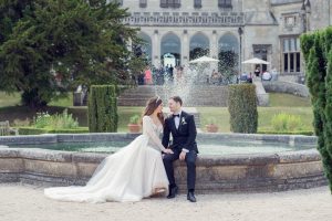 A bride in a white gown and a groom in a black tuxedo sit on the edge of a stone fountain, facing each other, with a grand castle and lush greenery in the background.