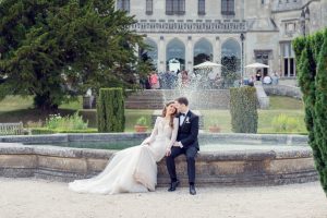 A bride and groom in formal wedding attire sit together by a stone fountain in front of a grand historic building, embracing and smiling, with guests visible in the background.