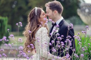 A bride and groom, dressed in a lace wedding dress and black tuxedo, smile and lean in close amid blooming purple flowers in an outdoor garden setting.