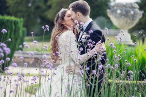 A bride and groom share a kiss outdoors, surrounded by tall purple flowers, with greenery and a stone urn visible in the background. The bride wears a white gown and tiara, and the groom is in a black suit.