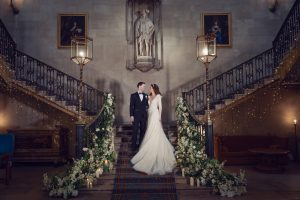 A bride and groom stand on a grand staircase decorated with flowers and candles, beneath a statue in an elegant hall with portraits on the walls and ornate railings, gazing at one another.