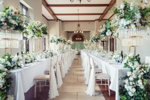 A beautifully decorated wedding banquet hall with long tables covered in white linen, elegant floral centrepieces, white chairs, and chandeliers hanging from the ceiling.