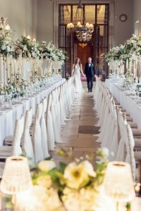 A bride and groom walk hand in hand down a long, elegantly decorated aisle lined with white chairs and tall floral centrepieces in a grand, chandelier-lit wedding hall.