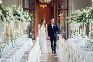 A bride and groom walk hand in hand between two elegant, flower-adorned banquet tables in a grand, light-filled hall, dressed in a white dress and black dinner suit, smiling and looking joyful.
