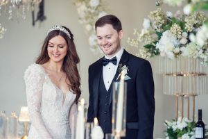 A bride in a white lace gown and a groom in a black tuxedo smile together indoors, surrounded by elegant white flowers and candles at their wedding.