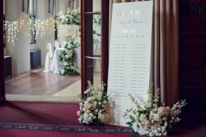 Large wedding seating plan with Take a Seat at the top, decorated with elegant floral arrangements at the base; fairy lights and round tables with white linen are visible in the background.