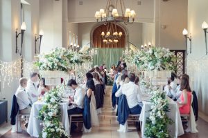 A wedding reception in a grand hall with long tables, elegant white floral centrepieces, and guests seated as a person gives a speech at the top table; fairy lights decorate the walls.