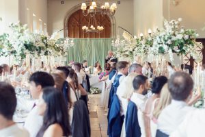 Wedding reception in a grand hall with guests raising glasses for a toast. Elegant floral centrepieces and chandeliers decorate the room. The top table, with the wedding party, is at the far end near green curtains.