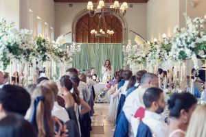 A bride stands and gives a speech at the top table during a wedding reception in an elegant hall, surrounded by seated guests and elaborate white floral centrepieces.
