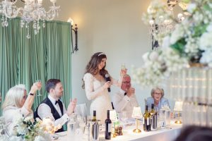 A bride in a white dress stands at a decorated table, holding a microphone and raising a glass for a toast. Guests next to her, dressed formally, also raise their glasses. The setting is elegant with chandeliers and flowers.