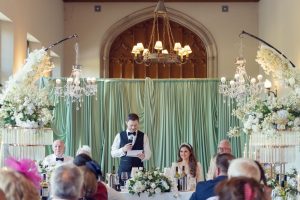 A man in a waistcoat stands at a decorated table, reading from paper at a wedding reception. Guests sit and listen, while the bride and others sit at the top table, surrounded by flowers and chandeliers.