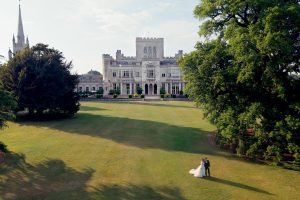 A bride and groom stand together on a large lawn, facing a grand historic mansion with towers and arched windows, surrounded by tall trees and gardens.