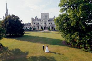 A bride and groom walk hand in hand across a large grassy lawn towards an elegant, historic mansion surrounded by tall trees, with a church spire visible in the background.