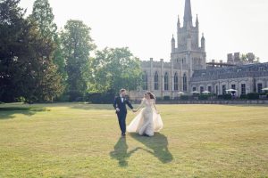 A bride and groom walk hand in hand across a sunlit lawn, smiling, with a large, ornate stone church or castle in the background, surrounded by green trees.
