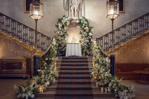A grand staircase decorated with lush white flowers and greenery, leading up to a small round table with a cake. Warm fairy lights and candles create an elegant, romantic atmosphere in a historic, ornate hall.