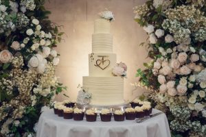 A tall, elegant white wedding cake decorated with flowers and a gold heart monogram stands on a round table, surrounded by cupcakes and lush floral arrangements of white and blush roses.