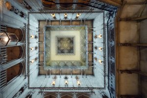 View looking directly up at an ornate, square, coffered ceiling with detailed carvings, surrounded by stone columns and walls, and illuminated by several golden wall sconces.
