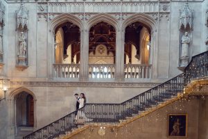 A couple in formal attire walks up an ornate stone staircase decorated with fairy lights inside a grand, historic building with arched windows, statues, and detailed carvings.