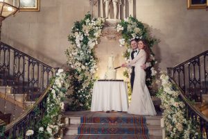 A bride and groom stand together on a grand staircase decorated with white flowers and greenery, smiling as they cut their wedding cake on a round table in front of them.