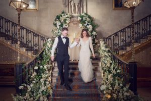 A bride and groom walk down a grand staircase decorated with white flowers and greenery. The bride wears a long white gown; the groom is in a black suit and bow tie. The setting is elegant and festive.