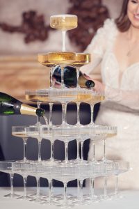 A bride in a white dress pours champagne into the top glass of a multi-level champagne tower, causing it to cascade into the glasses below. Another person is also pouring champagne from a bottle.