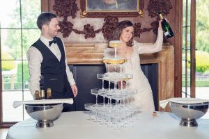 A bride in a white dress holds up a champagne bottle next to a groom in a waistcoat and bow tie. They stand by a table with a pyramid of stacked champagne glasses in an elegant, sunlit room.