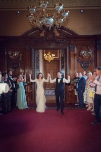 A newlywed couple holding hands and smiling as they enter a grand room with wood-panelled walls and chandeliers, while guests stand on either side applauding and celebrating.
