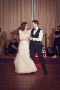 A bride in a white gown and a groom in a tuxedo dance together on a wooden floor, smiling at each other, with musicians and instruments visible in the background.
