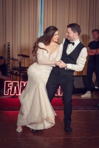 A bride in a white dress and a groom in a black dinner suit dance together, smiling and holding hands. In the background, musicians and illuminated “FAME” letters are visible.
