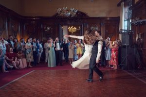 A bride and groom dance together on a wooden floor in an elegant, wood-panelled room, surrounded by smiling and clapping guests who watch and celebrate their first dance.