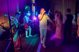 A band performs on stage with colourful lights while a singer in a sparkly shirt sings into a microphone; nearby, a bride and groom smile and dance with their guests.
