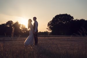 A bride and groom stand facing each other, holding hands in a sunlit field at sunset, with trees in the background and warm golden light surrounding them.