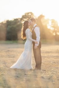 A bride and groom stand in a sunlit field, sharing a kiss. The bride wears a long white lace dress, and the groom is in a waistcoat and dress trousers. Soft golden sunlight creates a warm, romantic atmosphere.